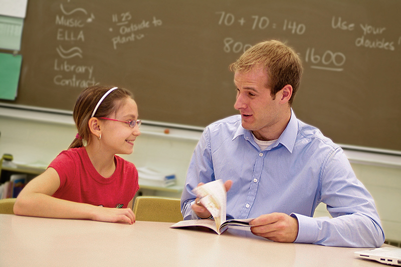 A young male teacher speaks to a female elementary school student at her desk